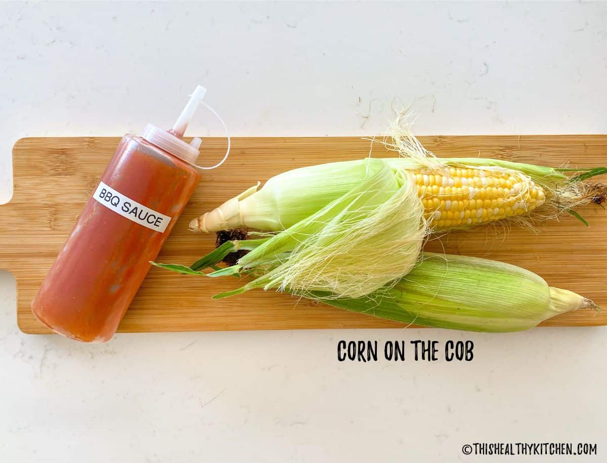 Corn on the cob on cutting board with bottle of bbq sauce beside it.