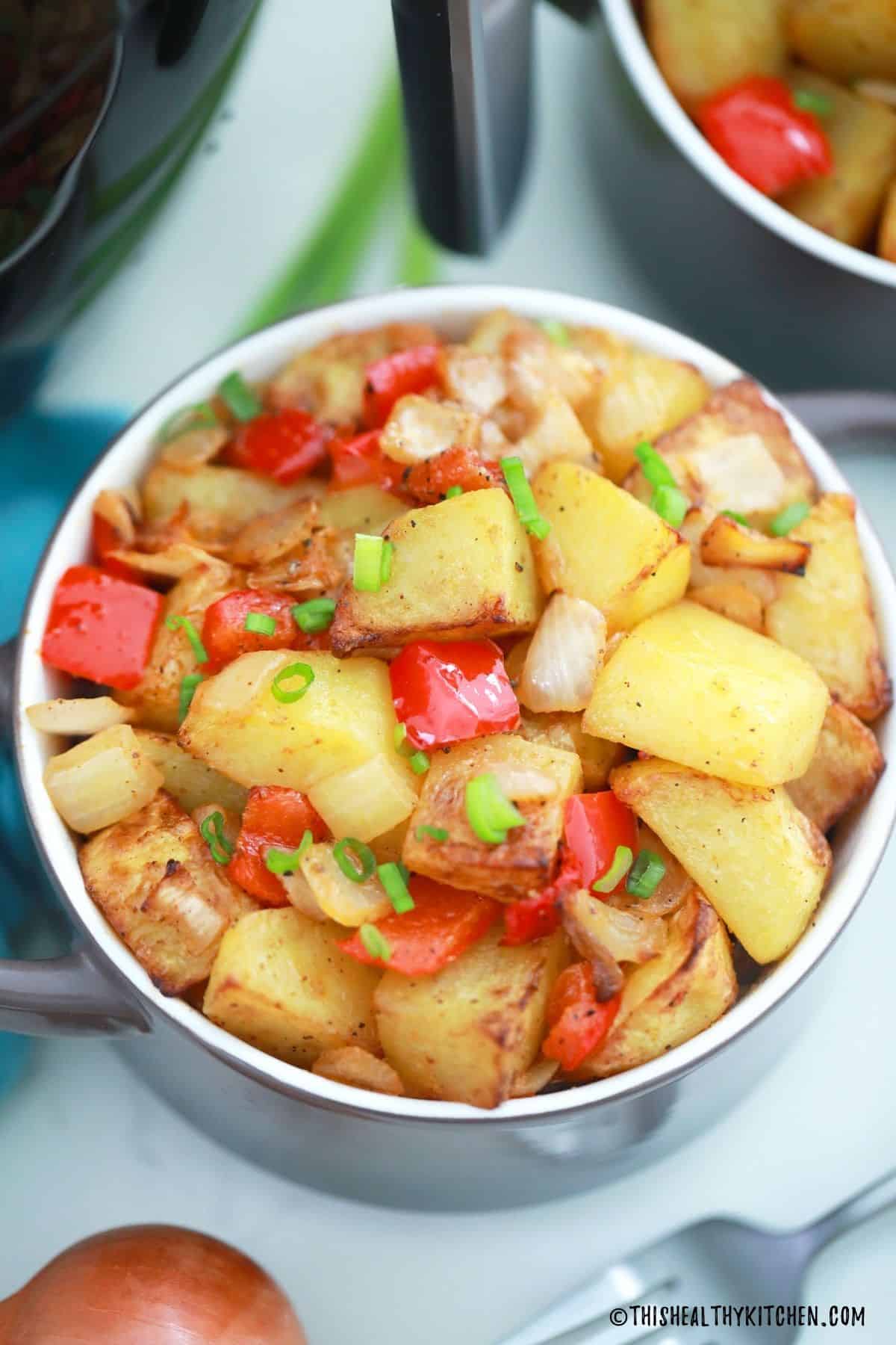 Close up of home fries in purple bowl with red pepper, onion and chives on top.
