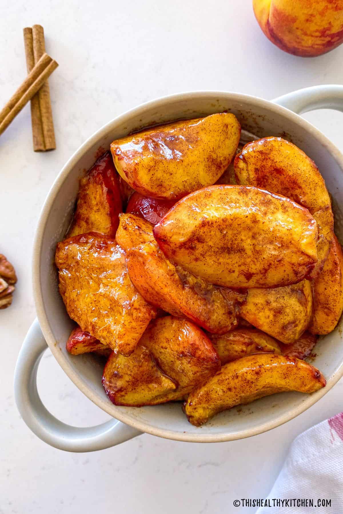 Air fried peaches in white serving dish with cinnamon sticks and pecans on the side.