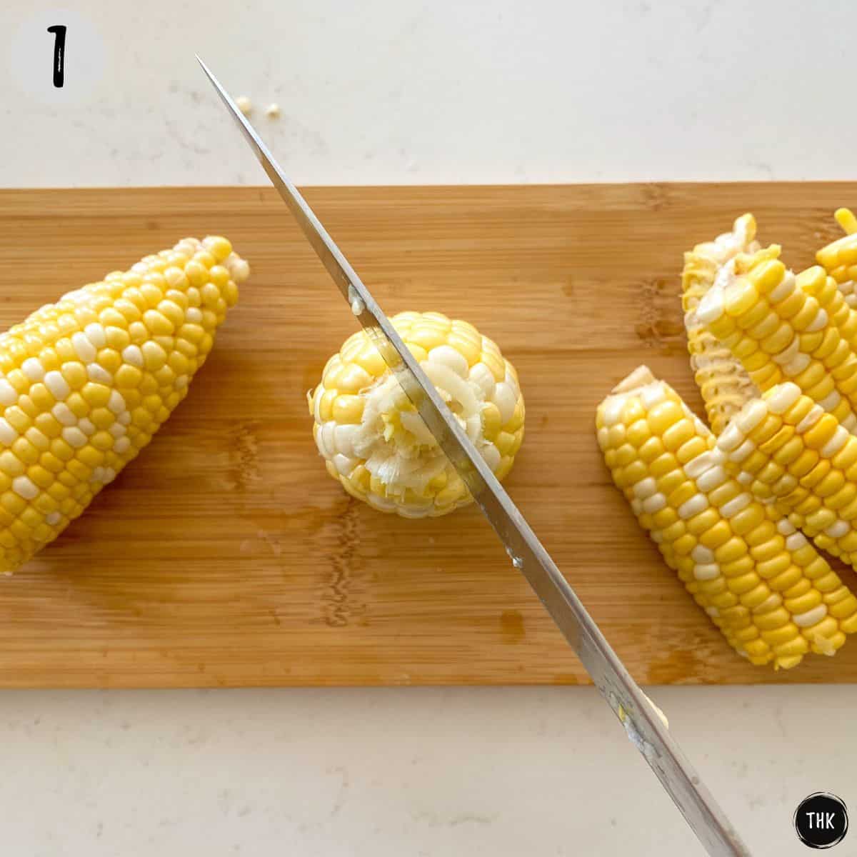 Halved corn sitting upright on cutting board with sharp knife about to cut it.