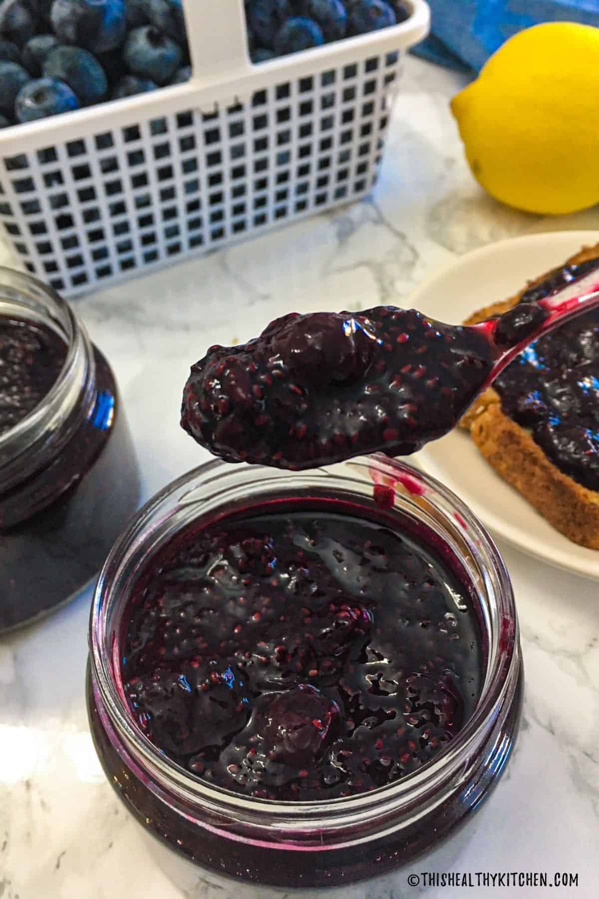 Spoonful of blueberry chia jam being held above jar of jam on kitchen counter.