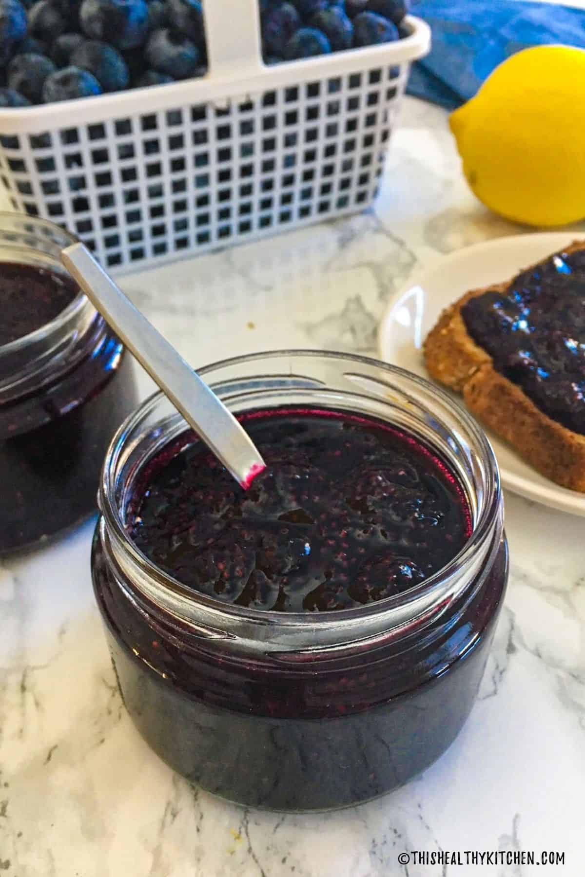 Jar of blueberry chia jam with spoon inside and blueberries in background.
