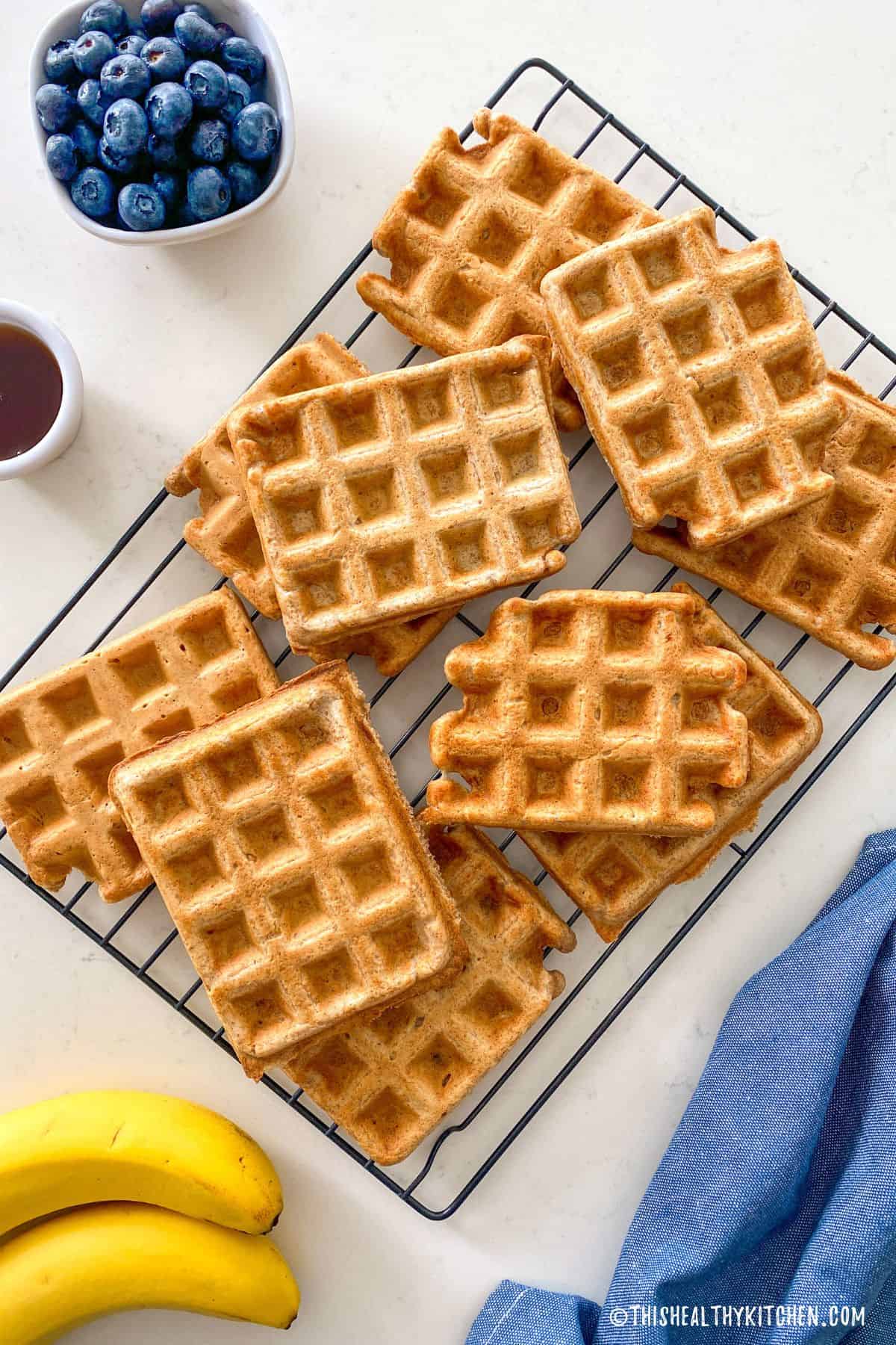 Cooling rack with 10 waffles on top and bowl of blueberries on the side.