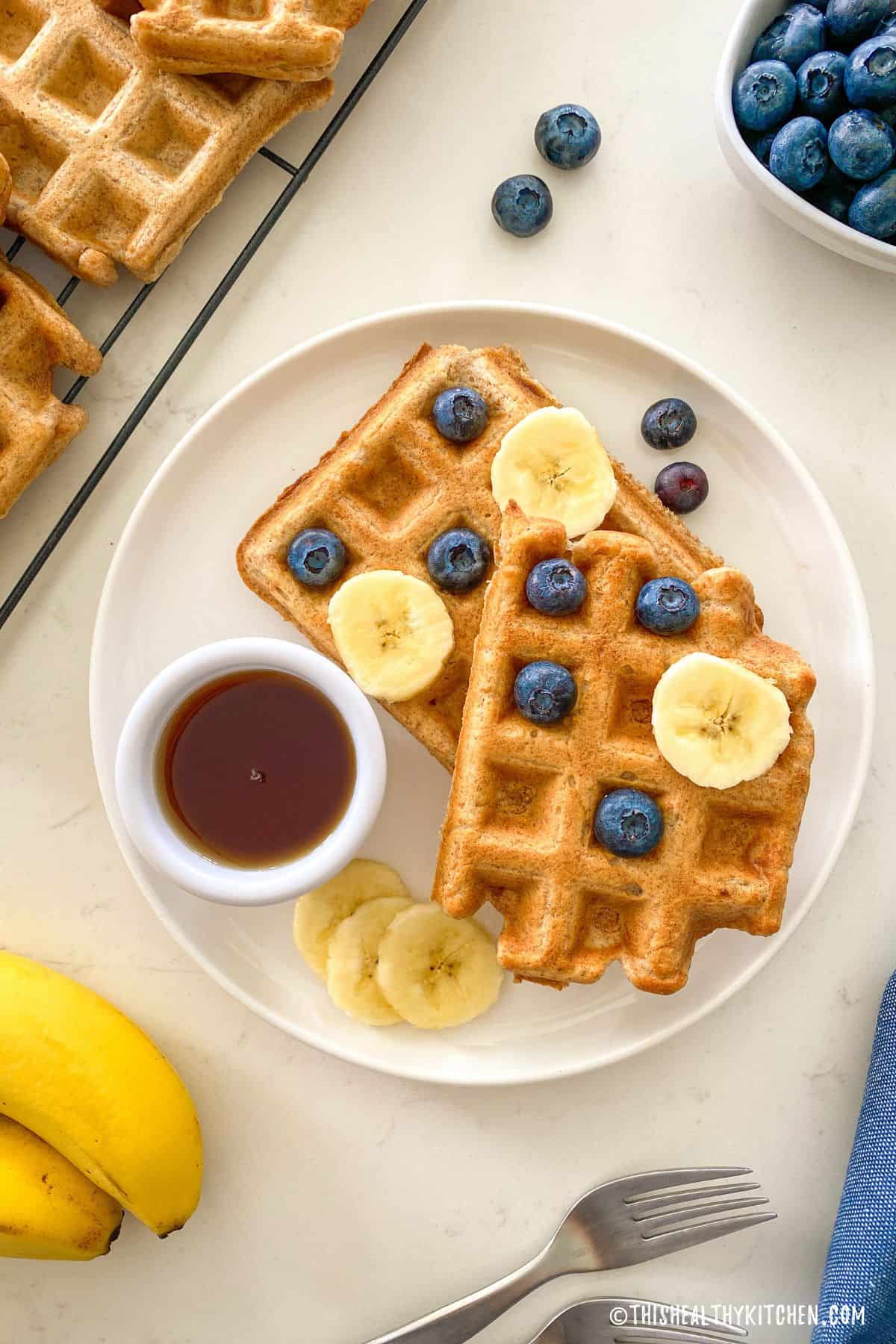 Plate with two waffles, with banana slices and blueberries on top and dipping bowl of maple syrup on the side.
