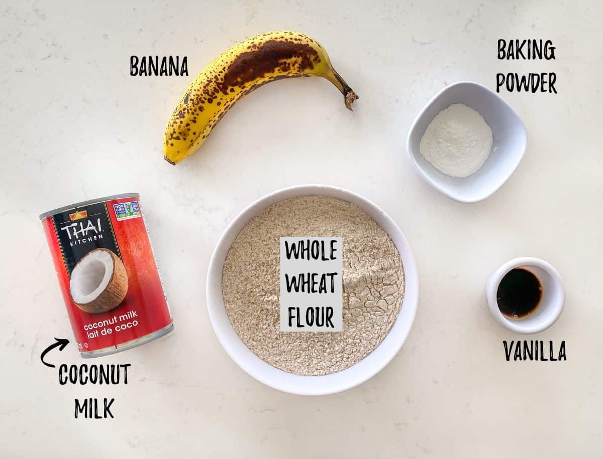 Bowl of flour, baking powder, vanilla, a banana and a can of coconut milk on kitchen counter.