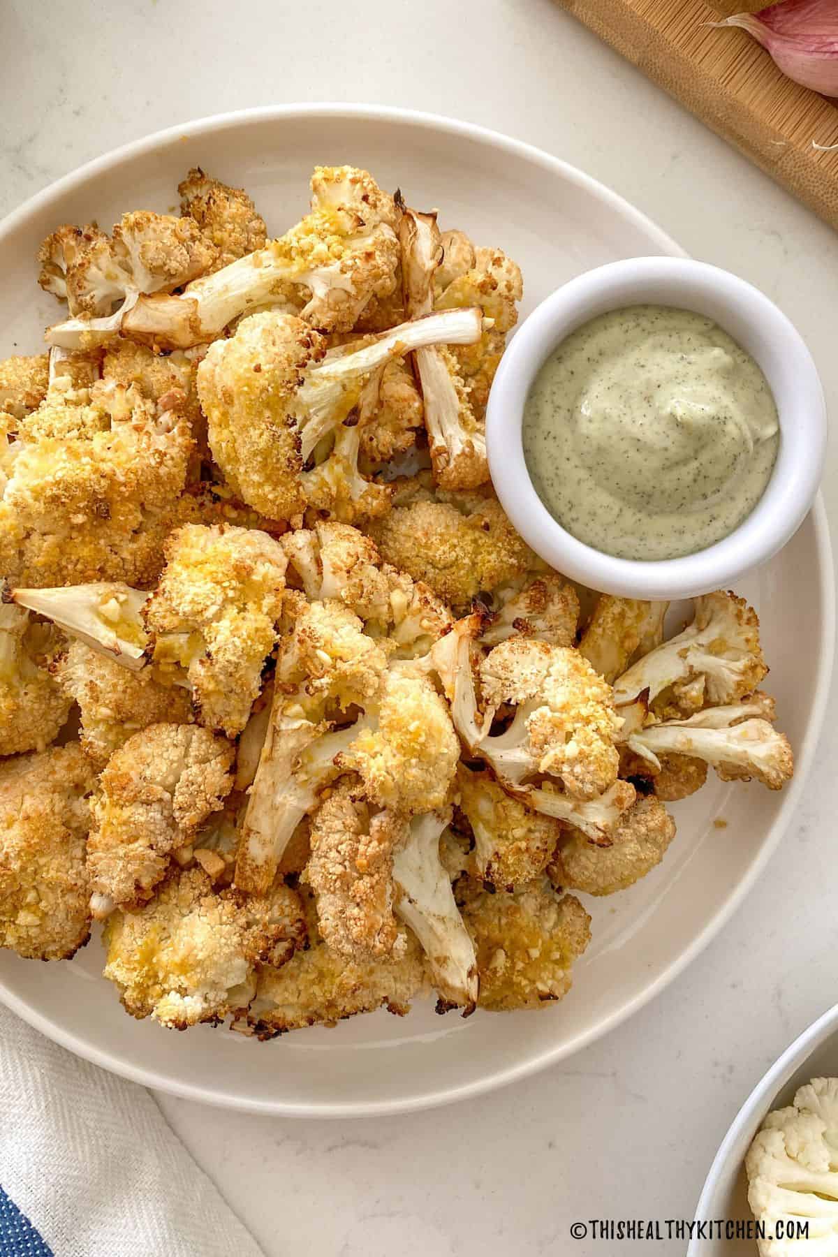 Roasted cauliflower in white plate with small bowl of dipping sauce beside it.