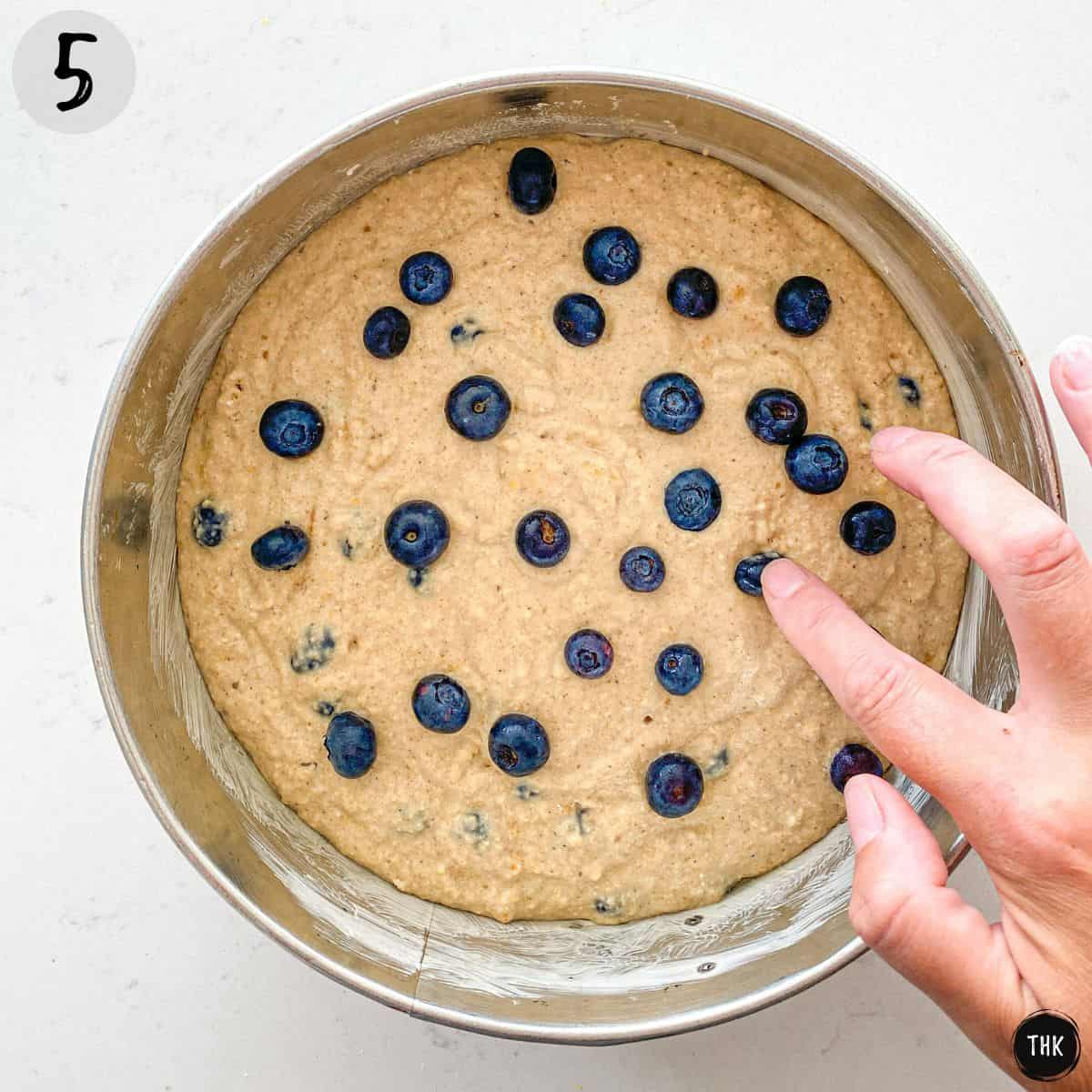 Cake batter in cake pan and finger poking blueberries inside it.