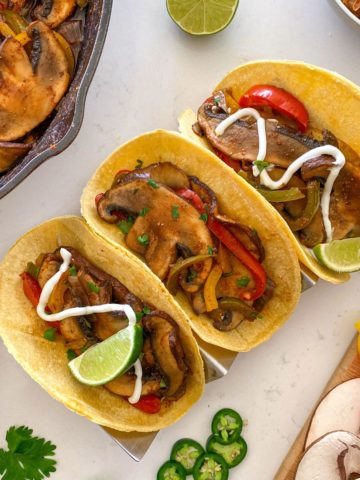 Three corn torillas filled with mushrooms, peppers, and sour cream and lime wedges on top.