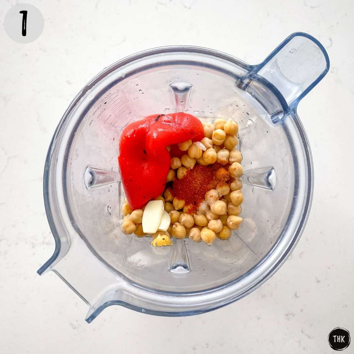 Blender with red peppers, chickpeas, garlic and seasoning inside.