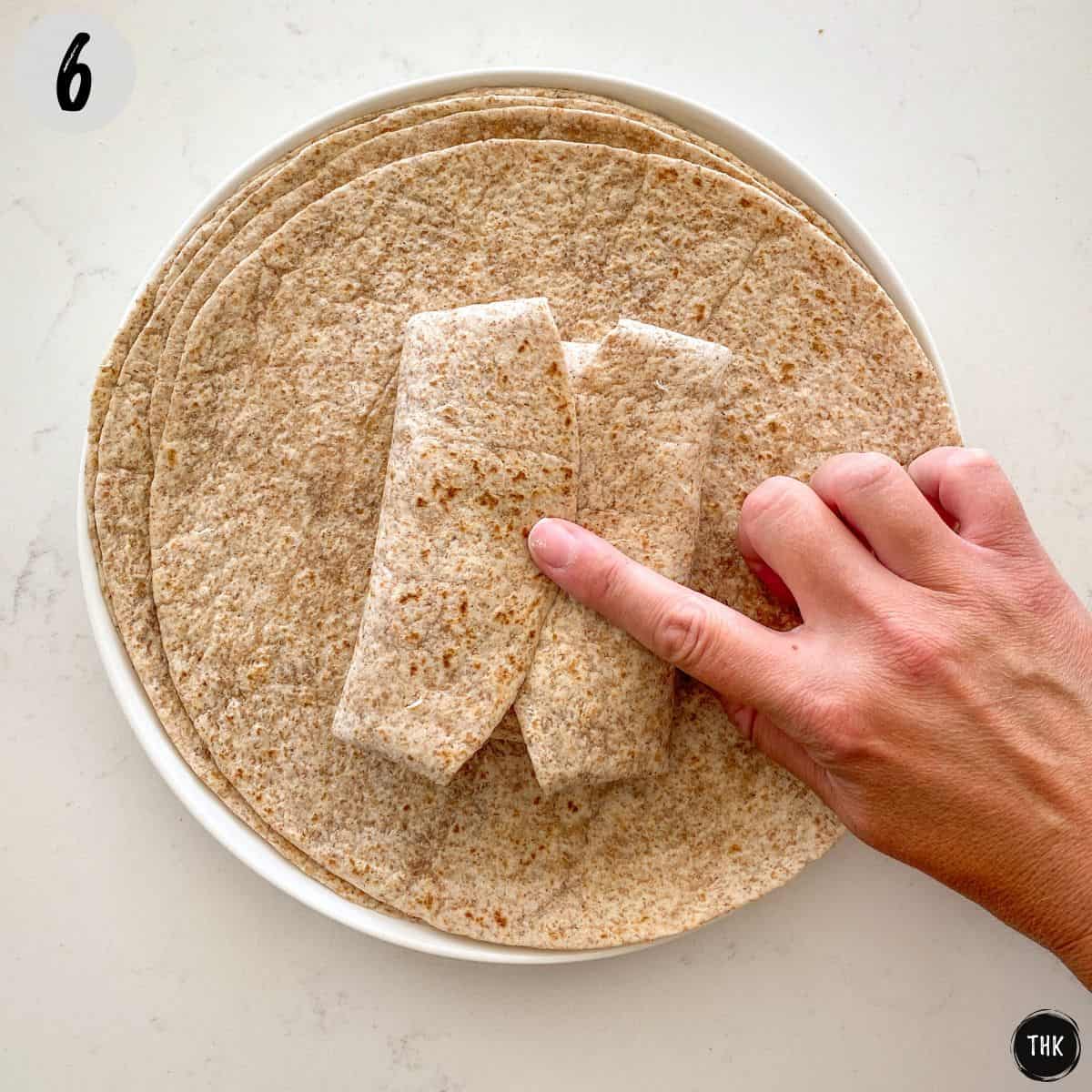 Hand holding closed a stuffed tortilla wrap on white plate.