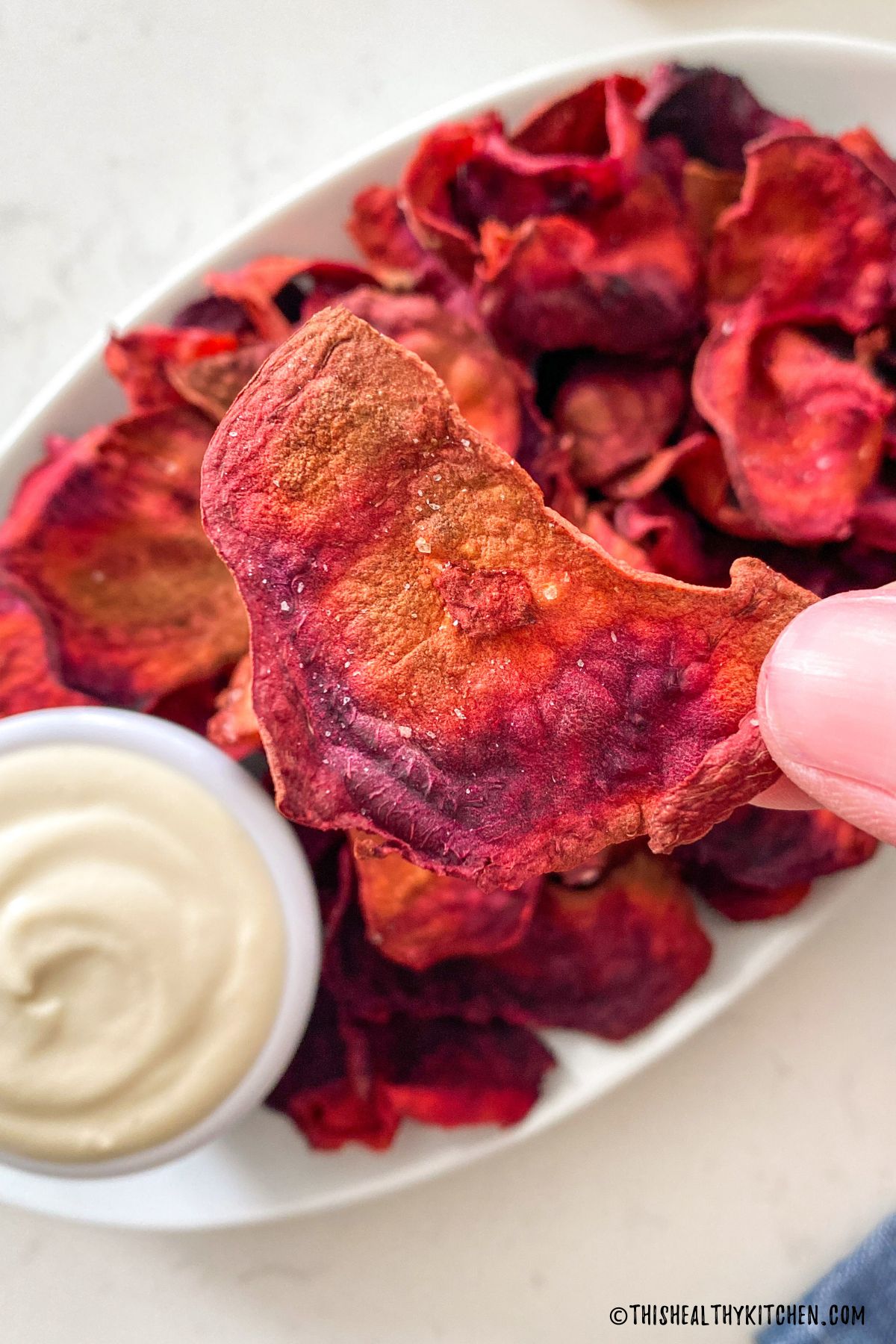 Hand holding up one beet chip with plate of chips below it.