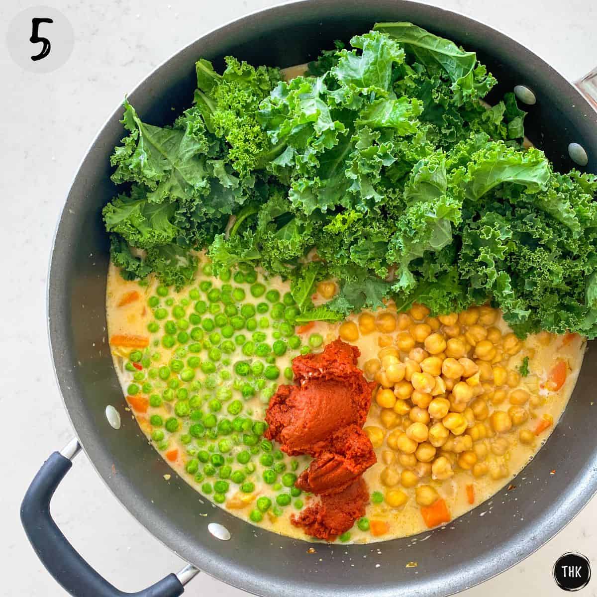 Deep skillet with chickpeas, red paste, peas, kale and creamy liquid.