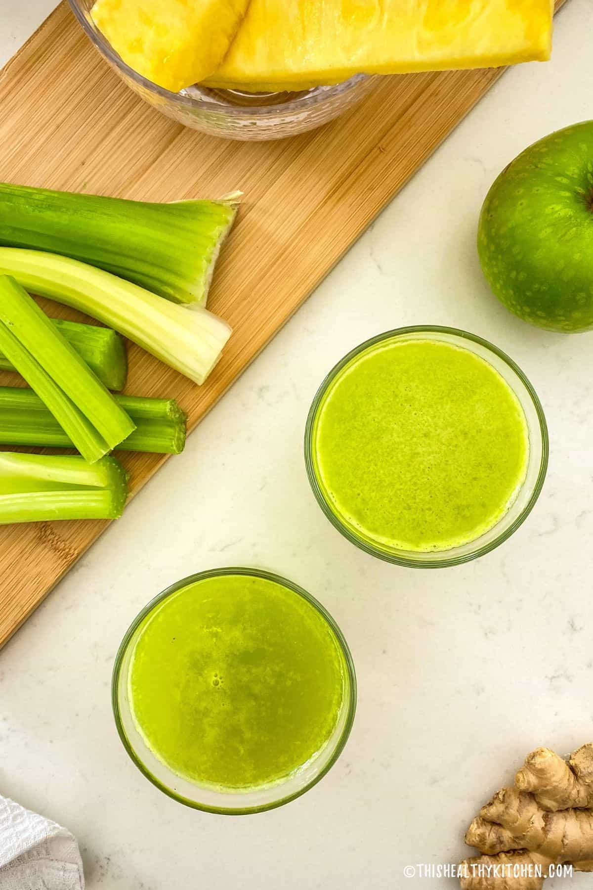 Two cups of green juice with celery, pineapple and green apple in the background.