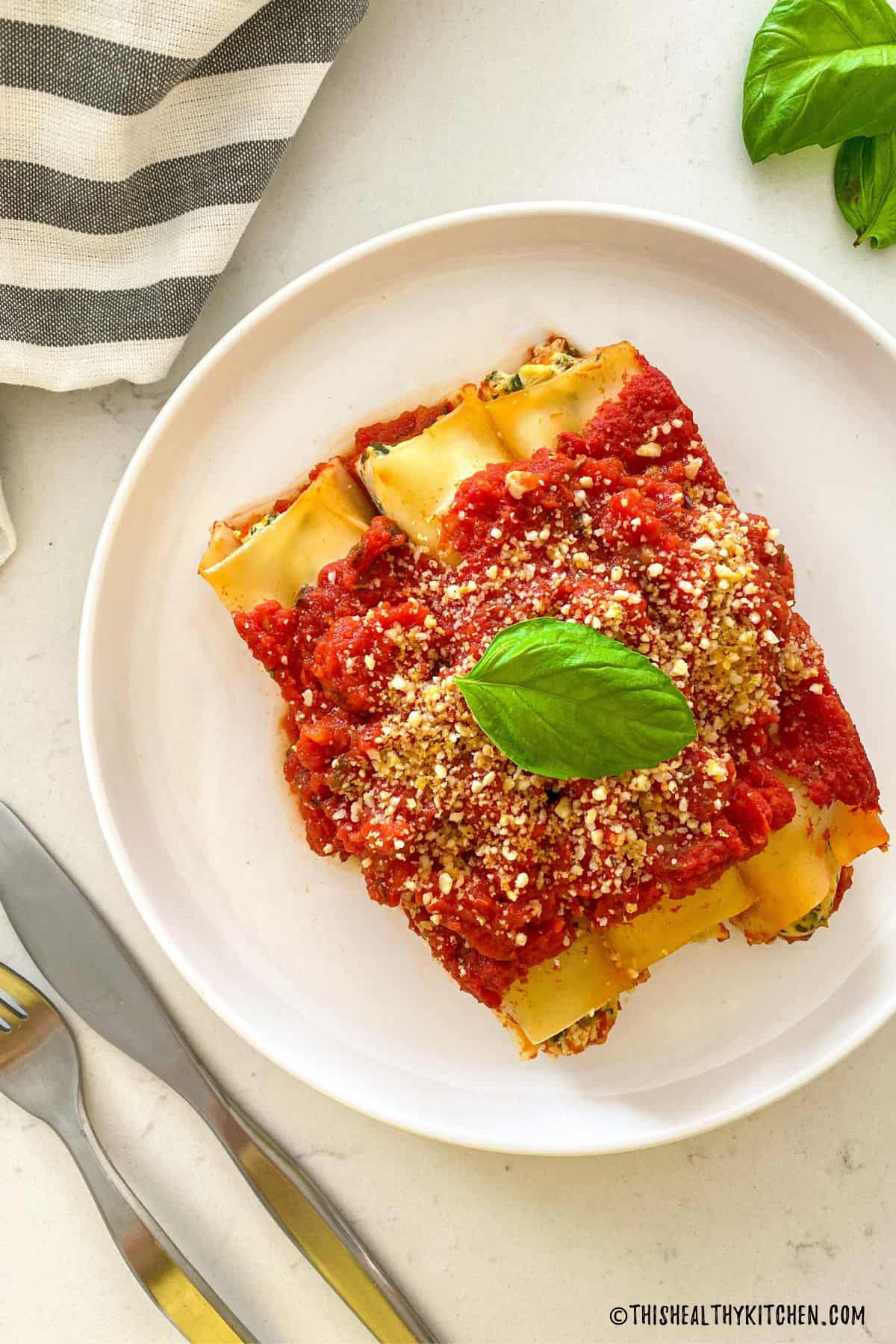 Three cannelloni in white plate with parmesan and fresh basil on top.