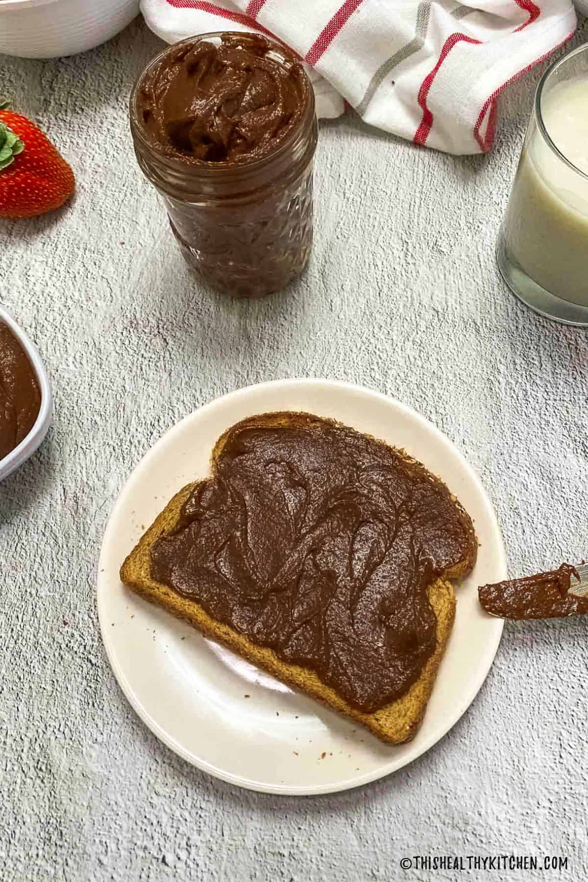Glass jar of chocolate spread behind toast on plate with chocolate spread.