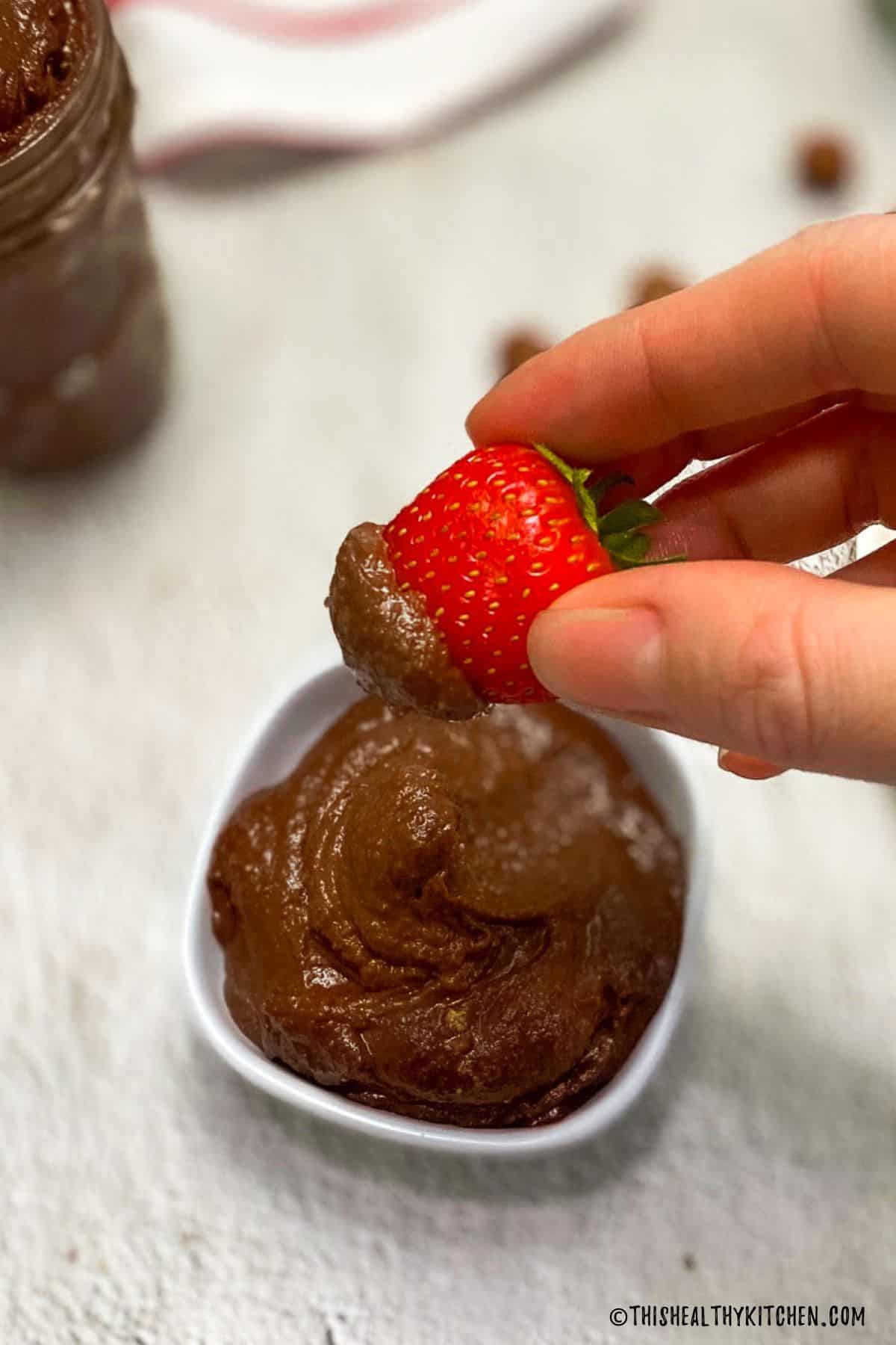 Strawberry dipped in chocolate spread being held up above bowl of Nutella.