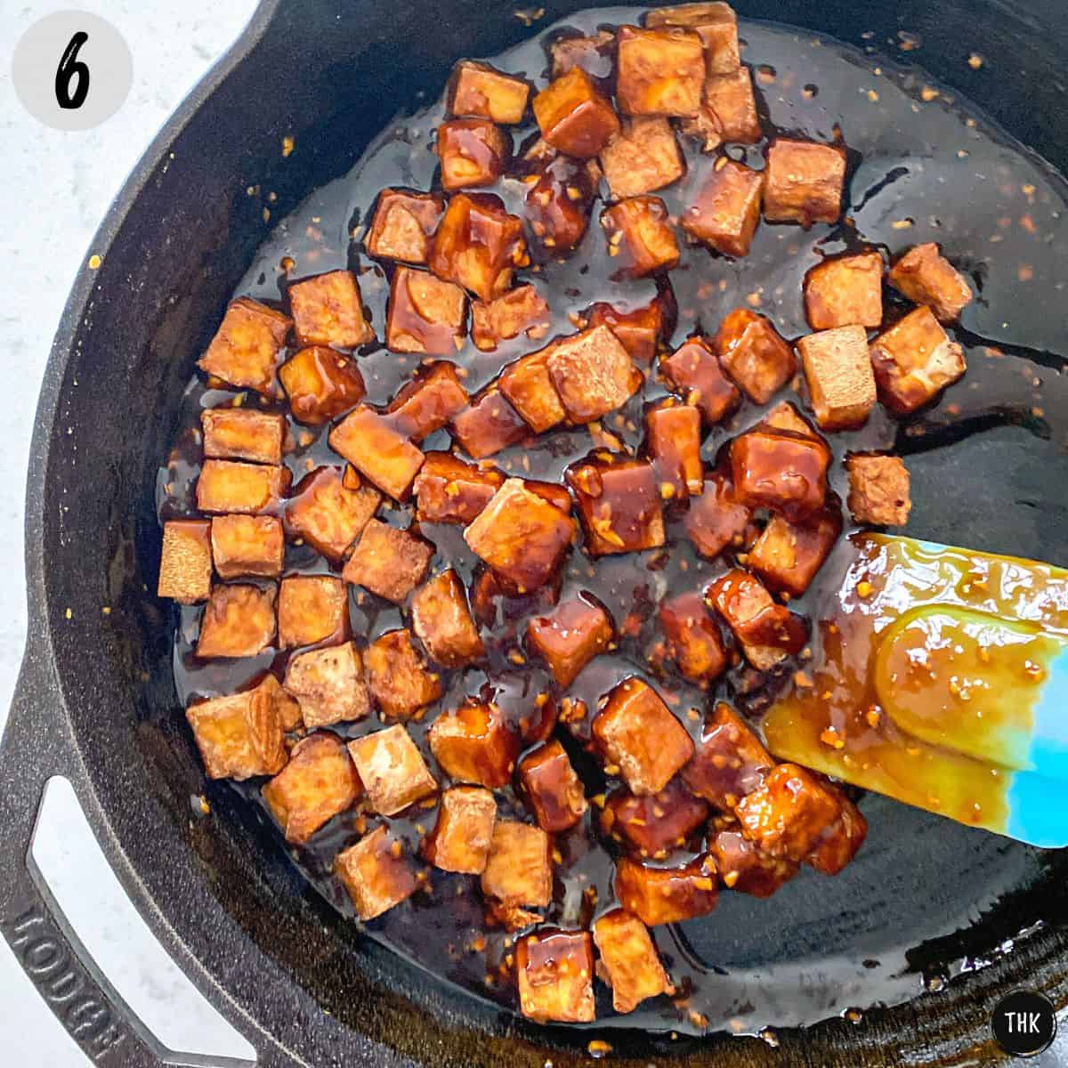 Tofu cubes inside cast iron pan being stirred into marinade.