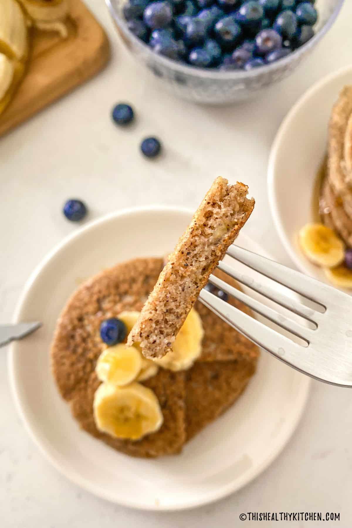 Bite of banana pancakes held up on fork with remaining pancake below.
