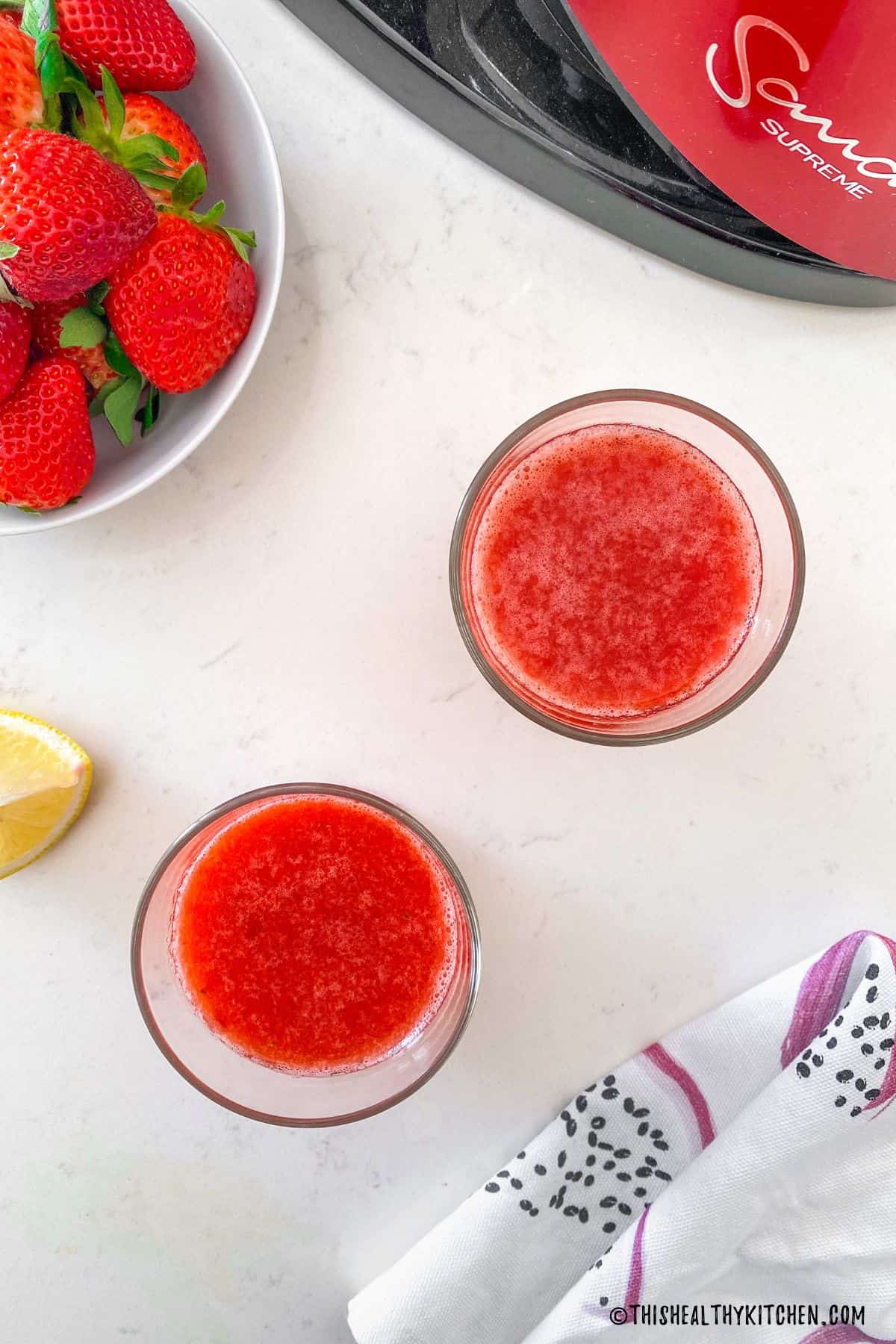 Overhead view of two glasses of red juice with bowl of strawberries beside them.