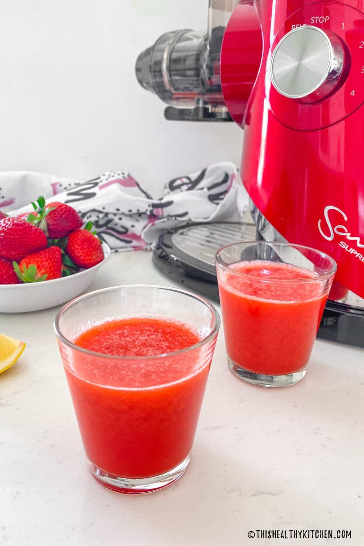 Two glasses of strawberry juice with juicer behind them.
