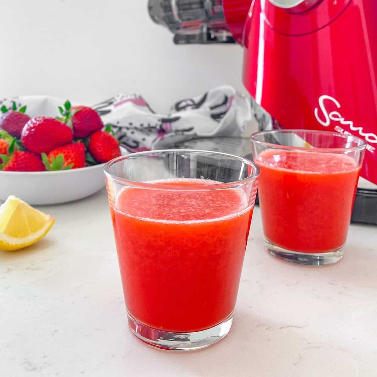 Two glasses of strawberry juice with juicer behind them.