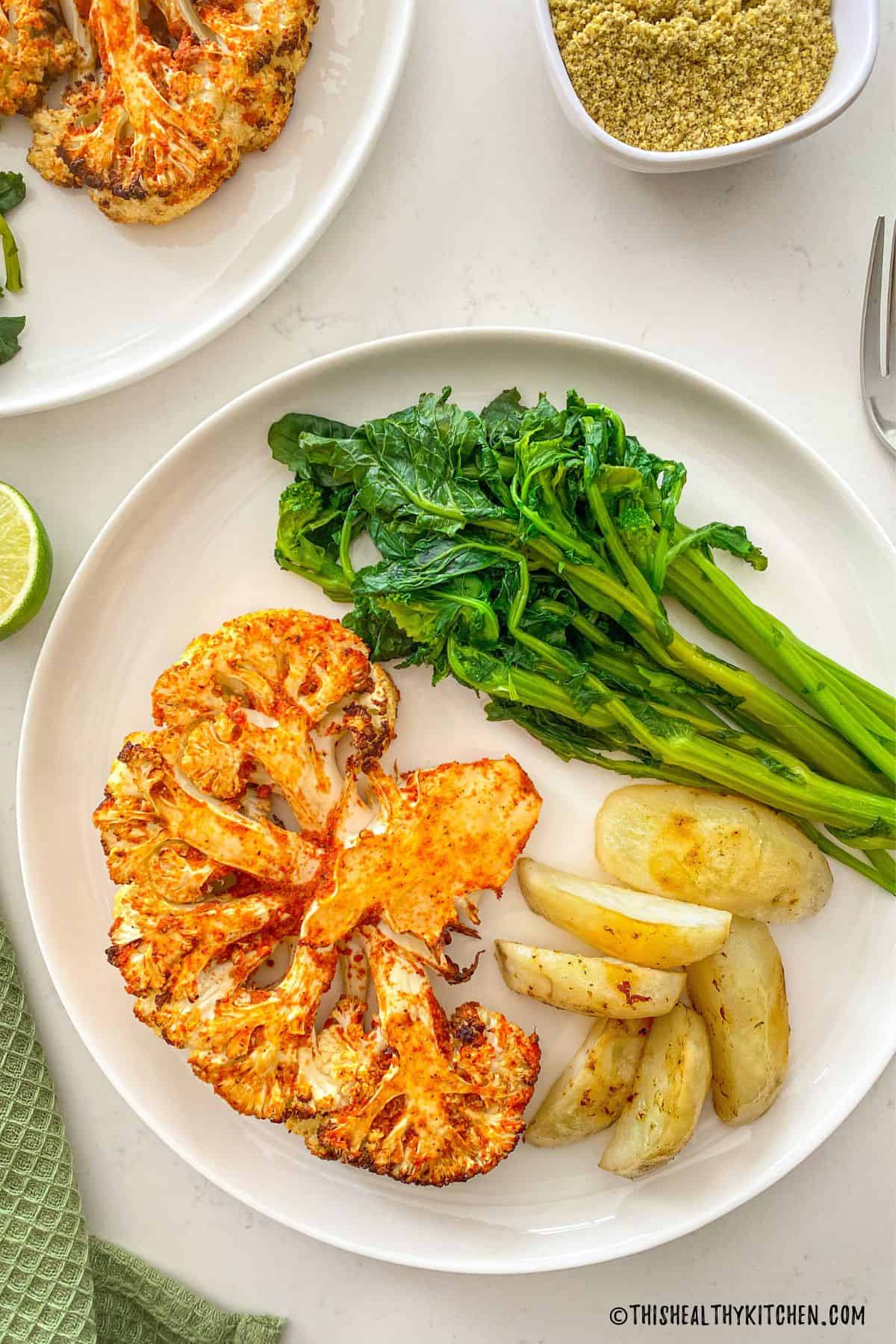 Air fried cauliflower steak on plate with potato wedges and rapini.