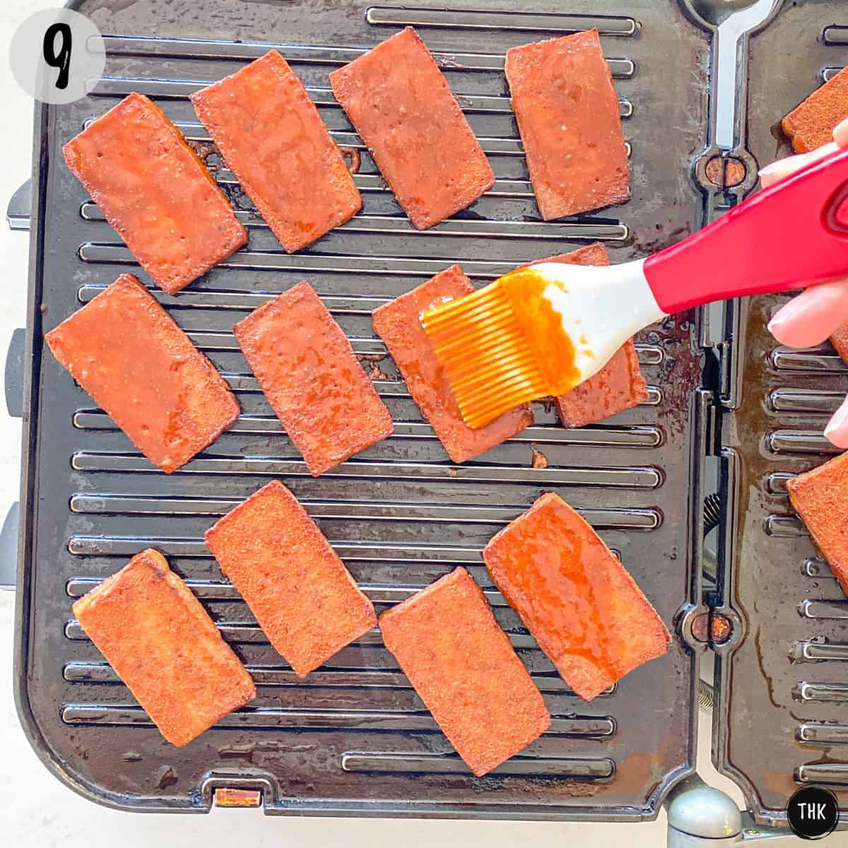 Smoked tofu slices on grill being brushed with red sauce.
