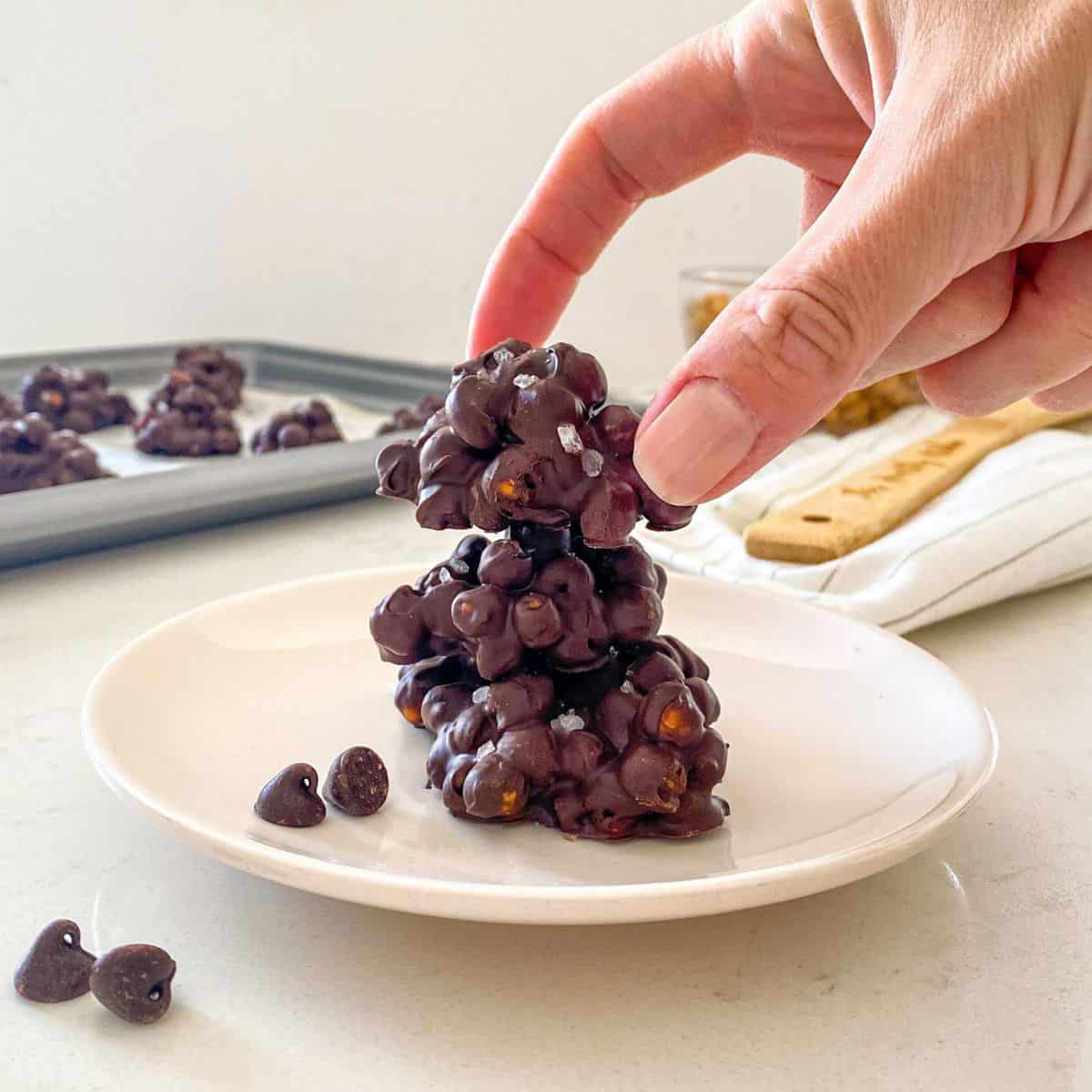 Chocolate chickpea clusters in a stack on a white plate with more in the background.