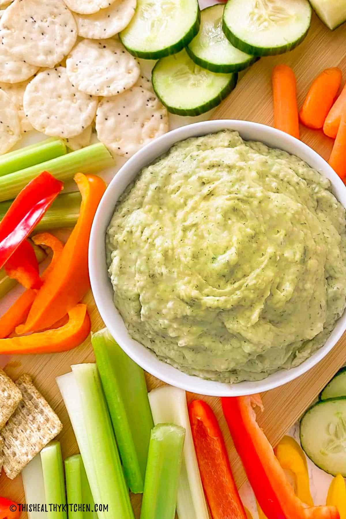 Bowl of green dip with veggies and crackers surrounding it.
