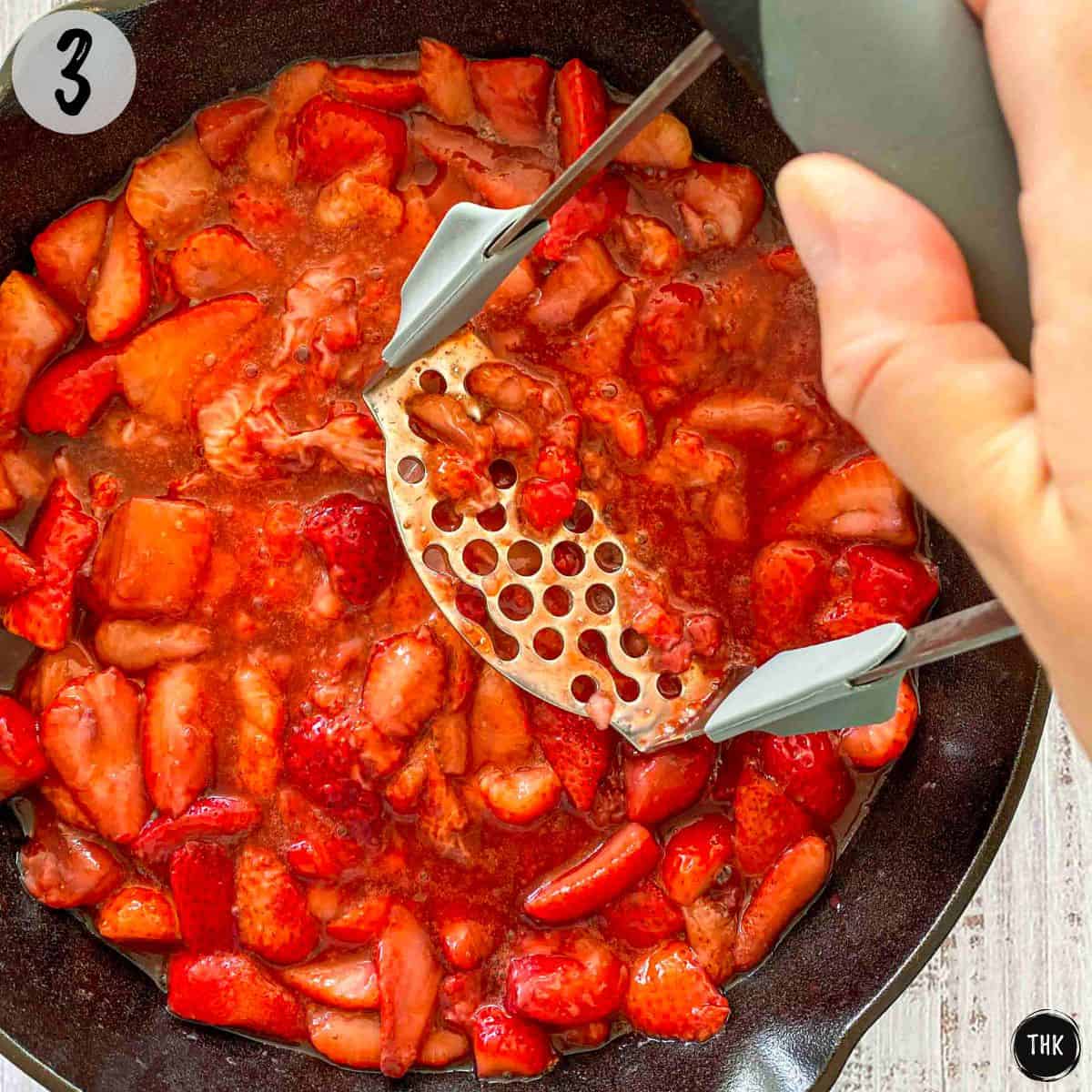 Strawberries being mashed with potato masher inside pan.