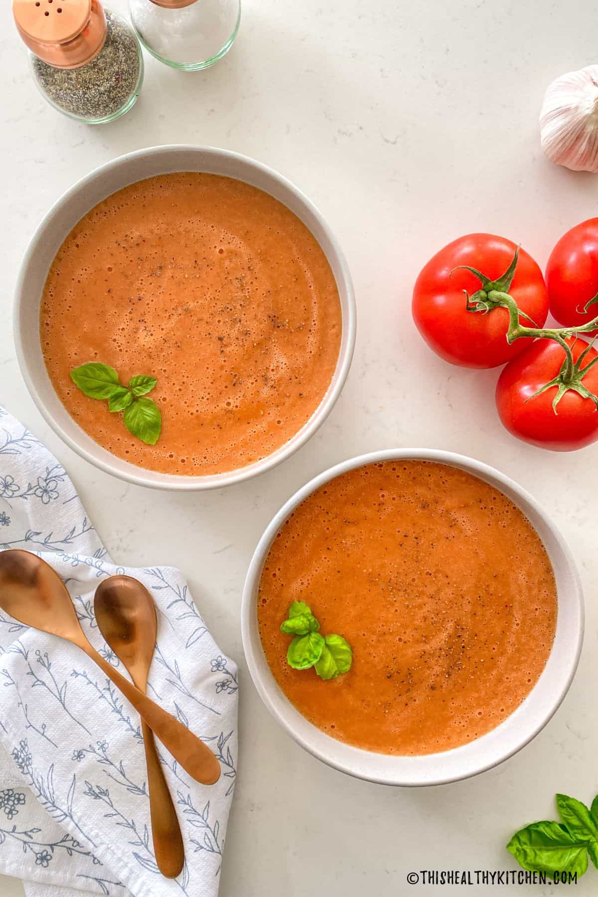 Two bowls of tomato soup with spoons beside them and basil leaves on top.