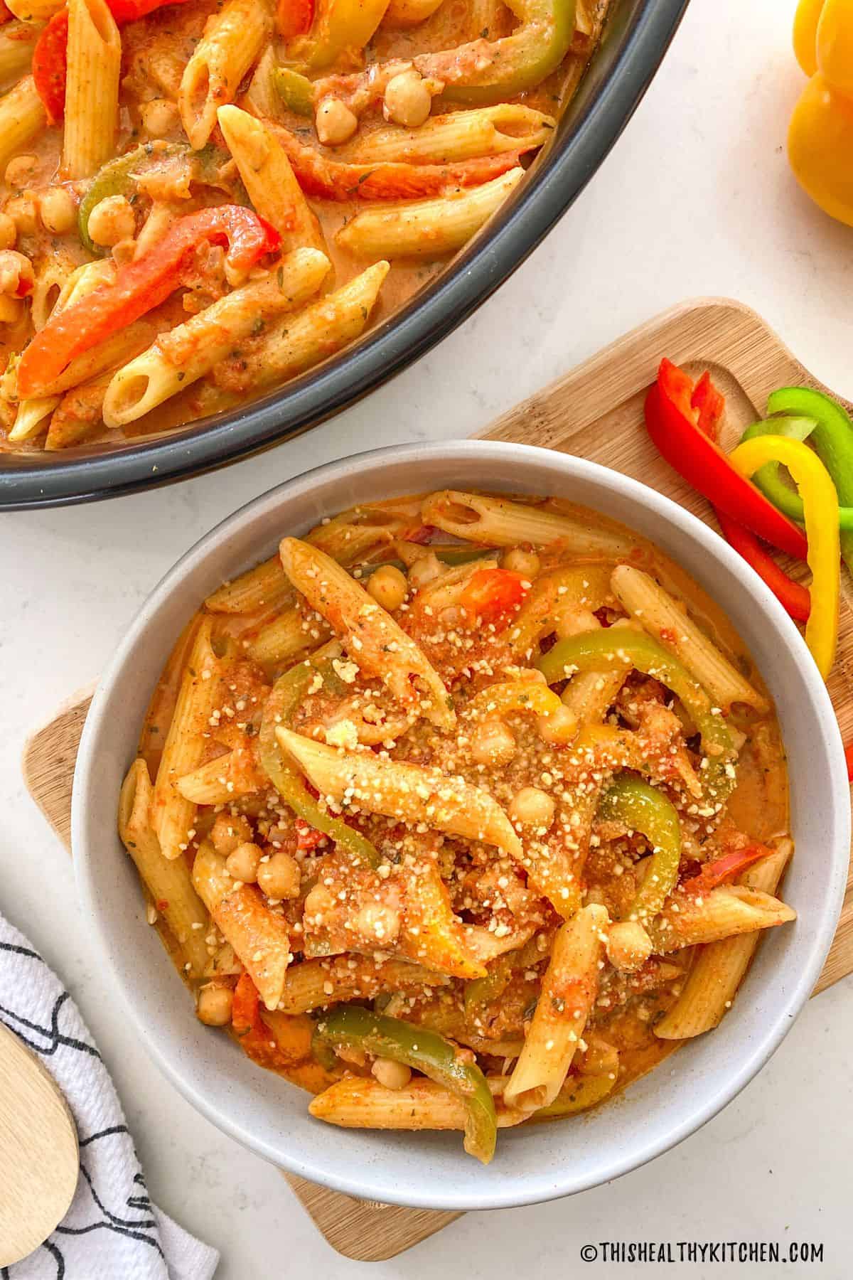 Vegan rasta pasta in skillet and a bowl with vegan parmesan cheese on top.