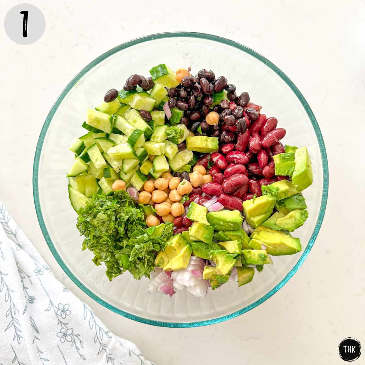 Mixing bowl with black beans, chickpeas, kidney beans, cucumber, avocado, onion, and basil.