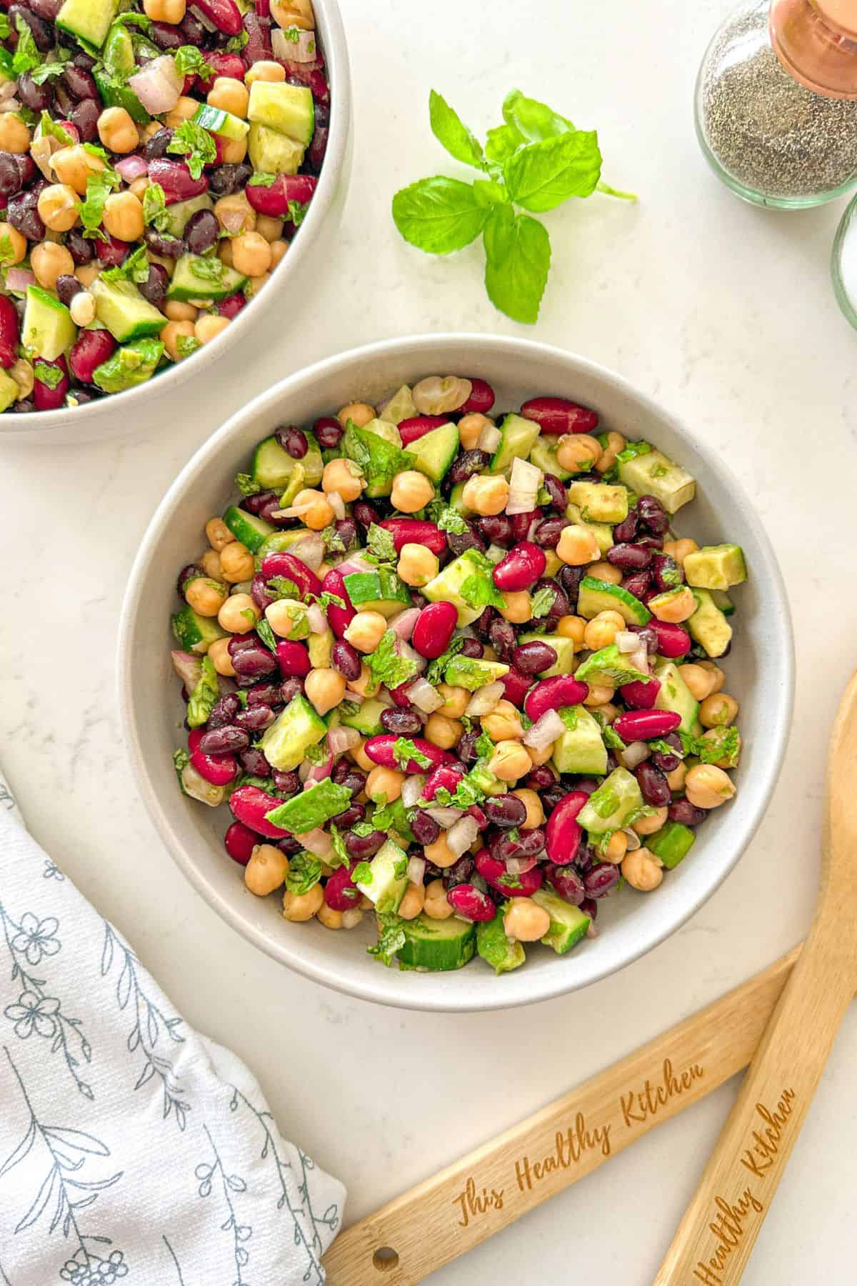 Two bowls of bean salad with cucumber, avocado, red onion, and basil.
