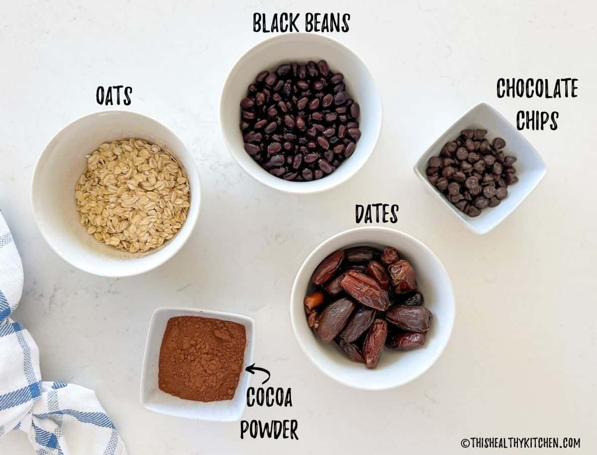 Bowls of black beans, oats, dates, cocoa powder, and chocolate chips on kitchen counter.