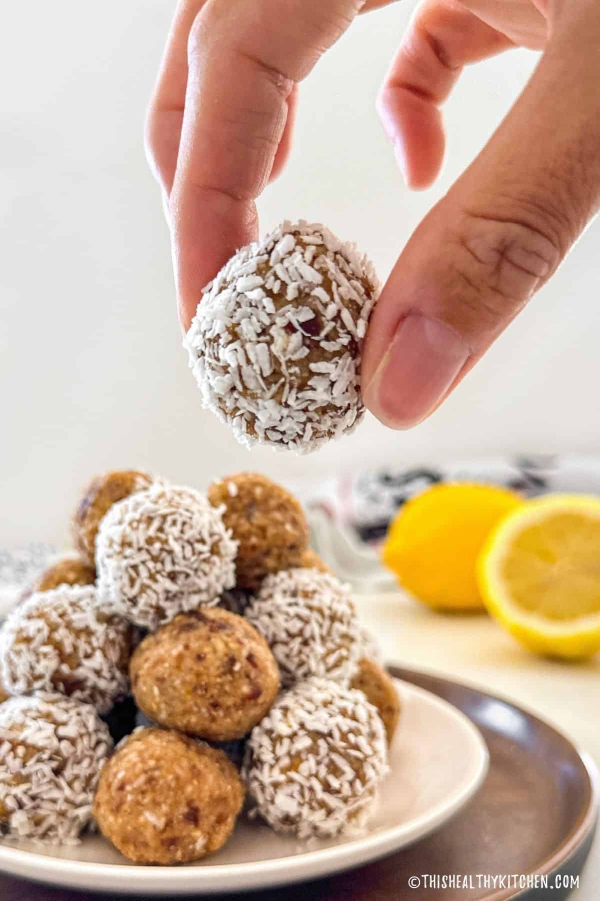 Hand holding up an energy ball with coconut around it and plate with more balls below.