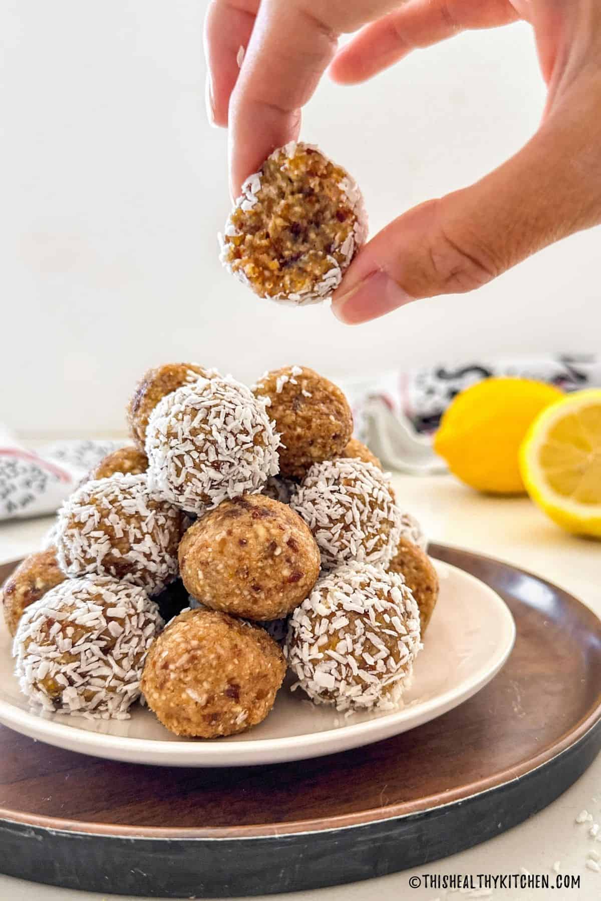 Plate of lemon bliss balls with hand holding up one that is bitten in half.