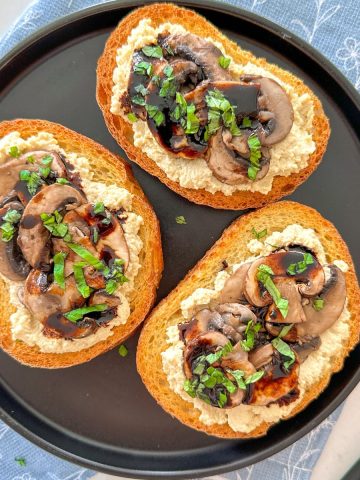 Three crostini on a black plate with vegan ricotta, sautéed mushrooms, and balsamic glaze on top.