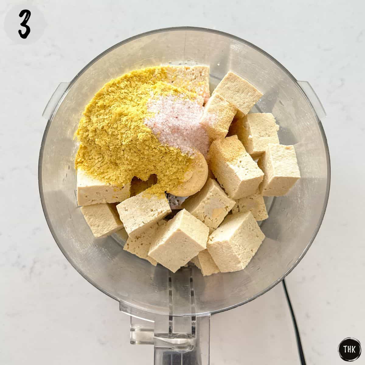 Tofu pieces, nutritional yeast, vinegar, and spices inside food processor.