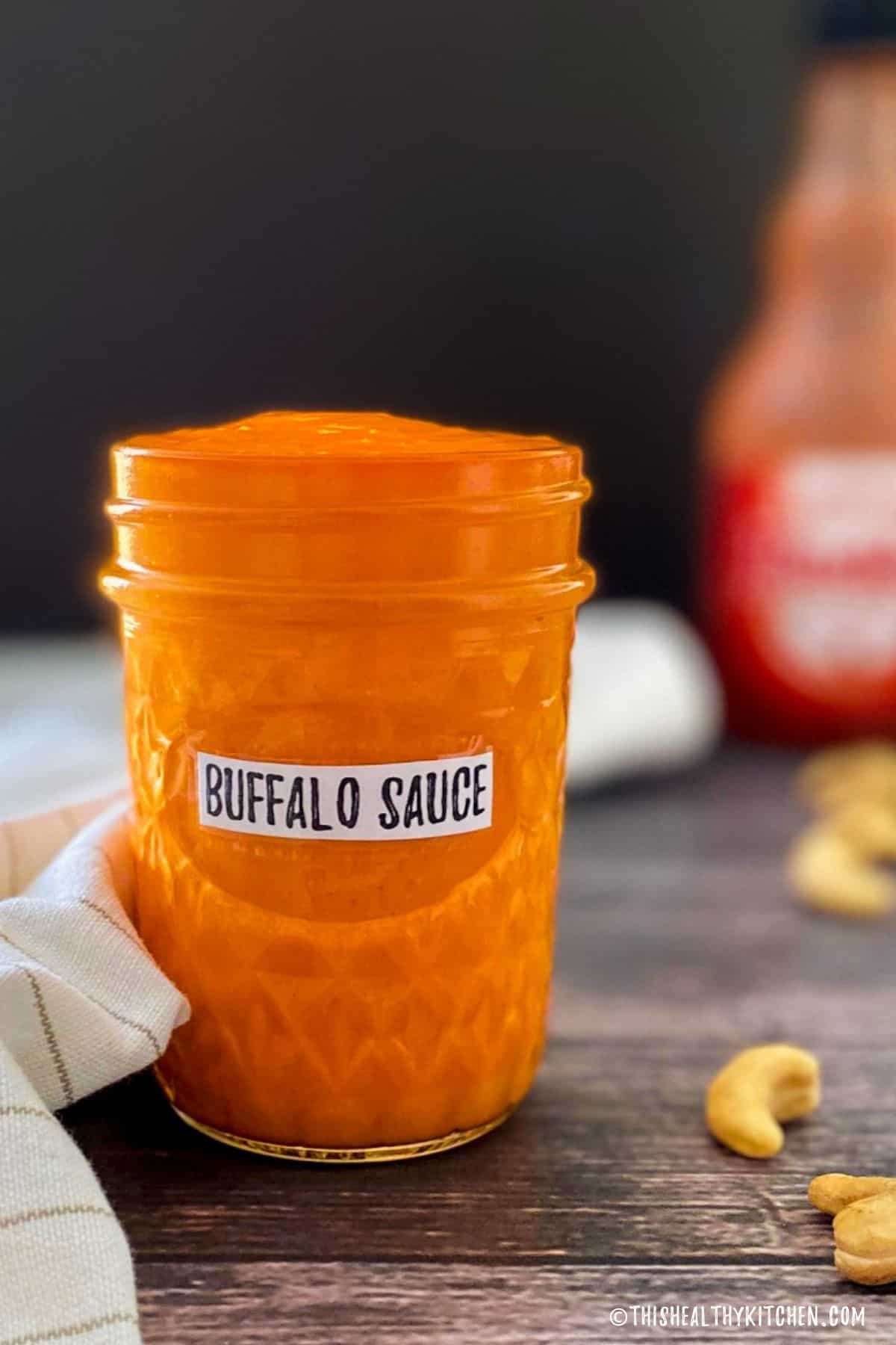 Glass jar with buffalo sauce inside and bottle of Franks hot sauce behind it.