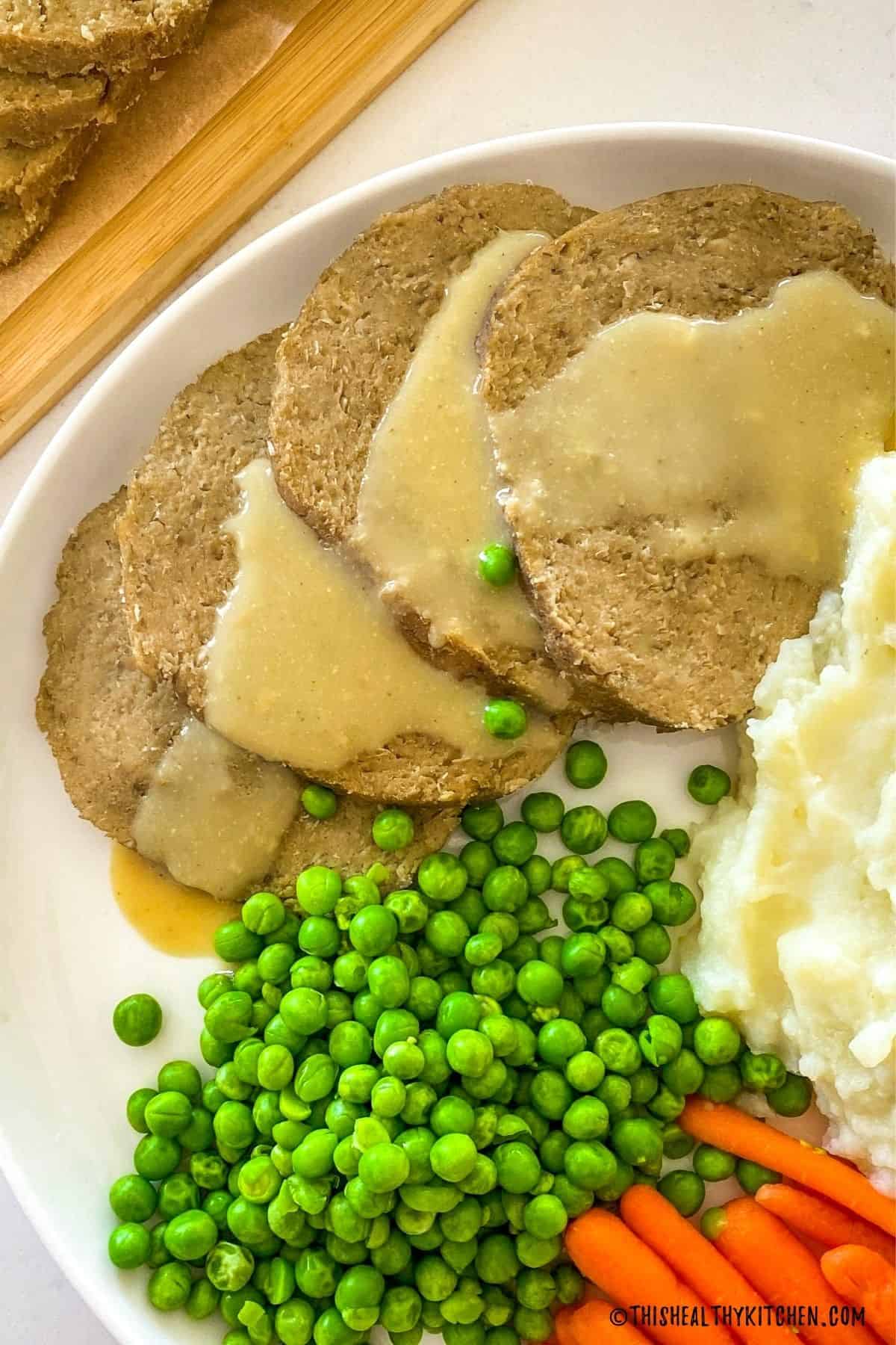 Sliced vegan roast on a plate with mashed potatoes, peas and carrots.