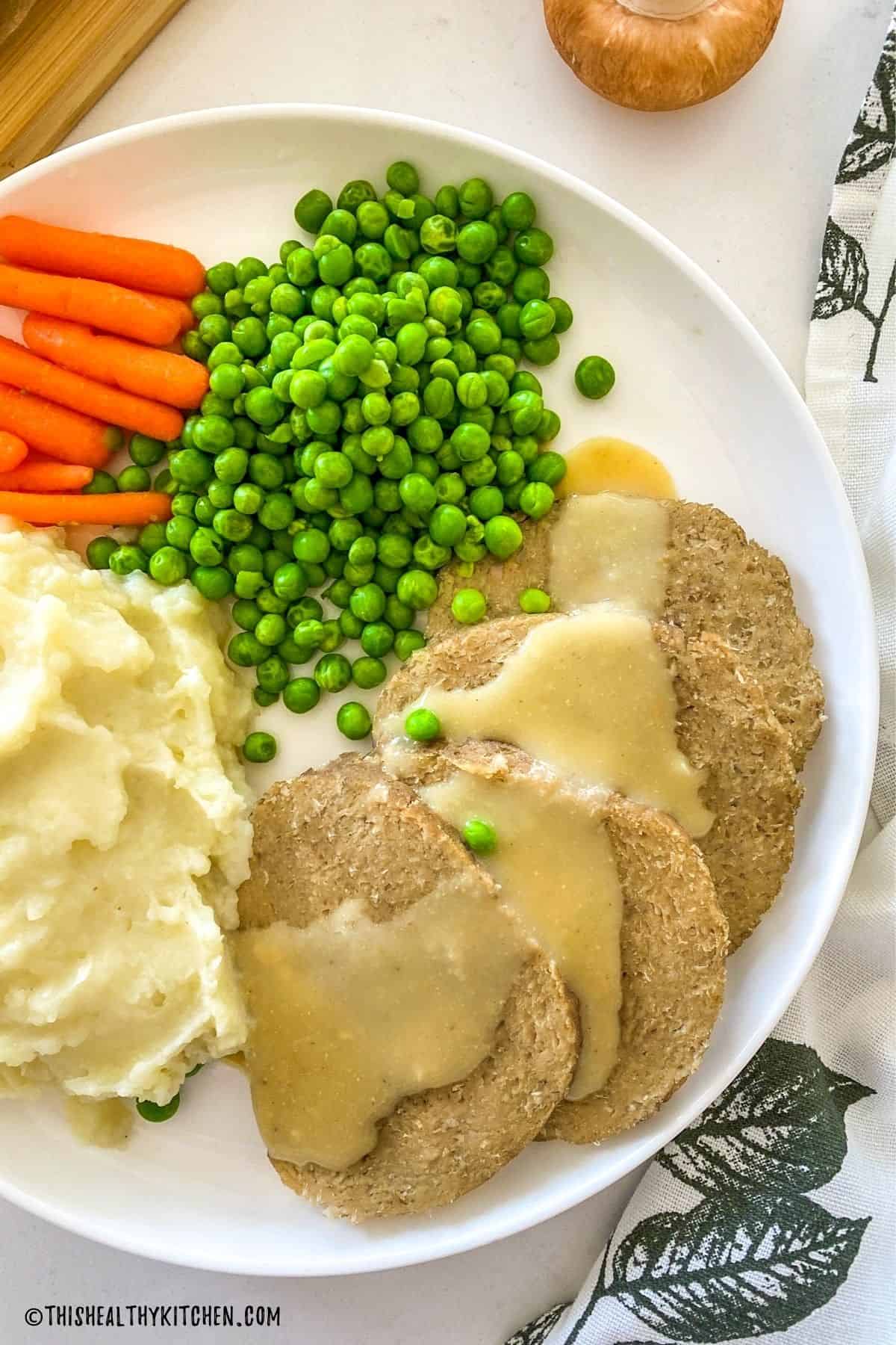 Four slices of seitan roast on a plate with gravy on top and veggies on the side.
