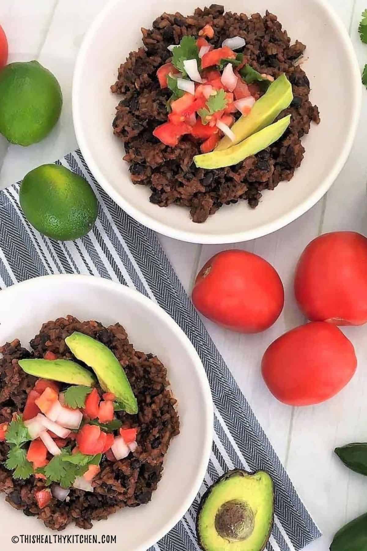 Two bowls of rice and beans with veggies and avocado slices on top.