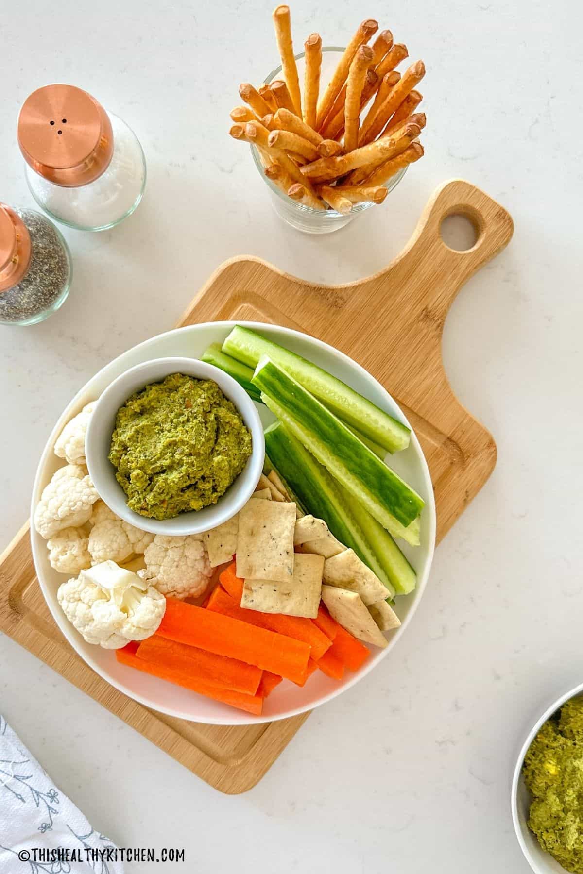 Small dipping bowl of pistachio pesto in plate with raw veggies and crackers around it.