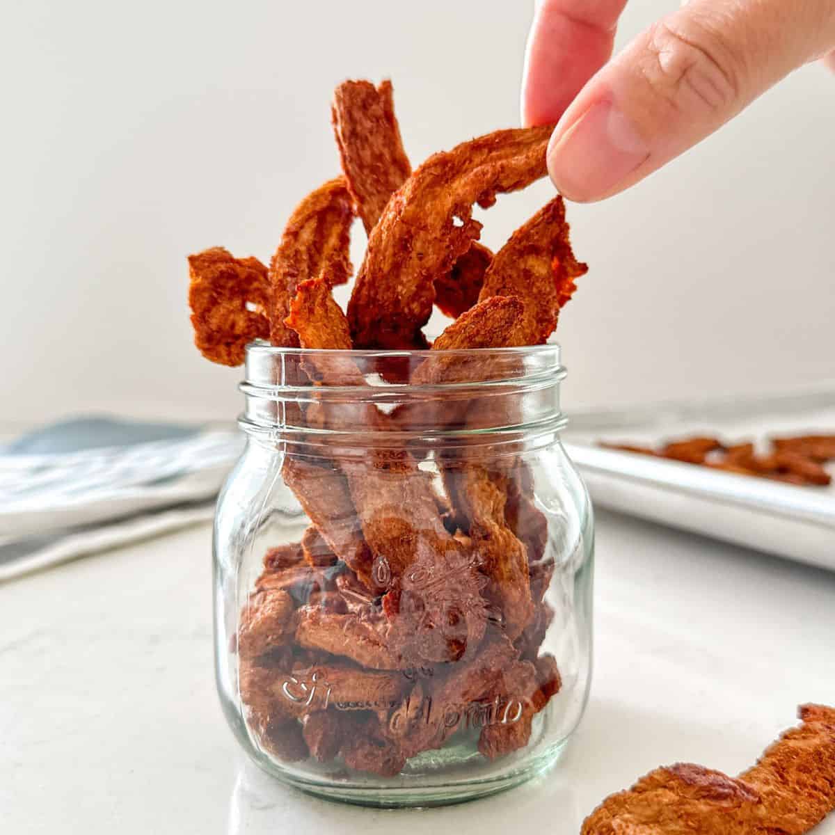 Hand pulling out a piece of vegan jerky from glass jar.
