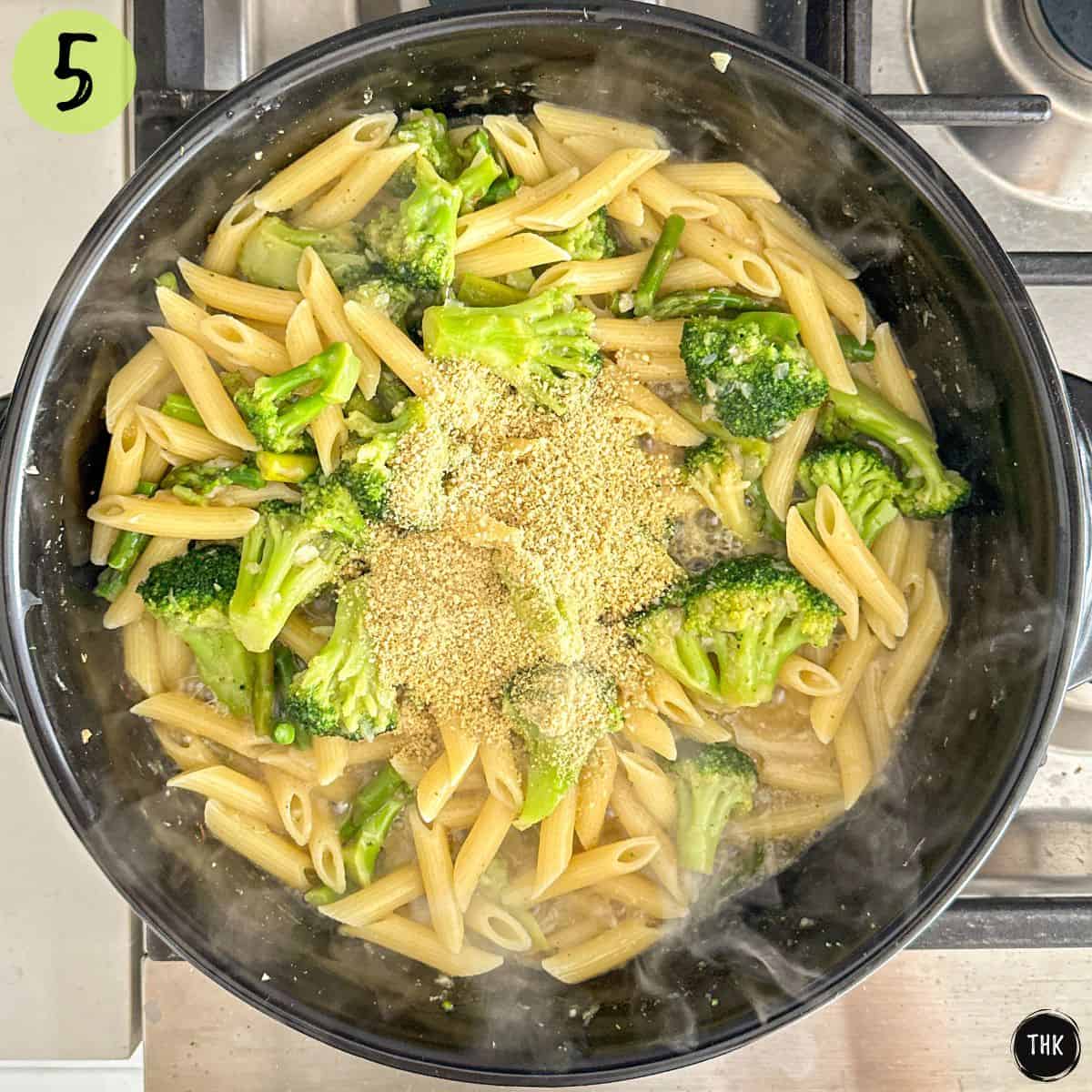 Broccoli, asparagus, penne pasta and vegan parmesan inside large skillet on stovetop.