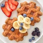 White plate with two banana protein waffles on it, with sliced bananas, strawberries and blueberries on top.