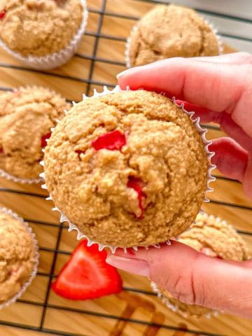 Hand holding up strawberry muffin above wire rack with more muffins.