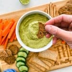 Cracker being dipped into bowl of green dip on cutting board.