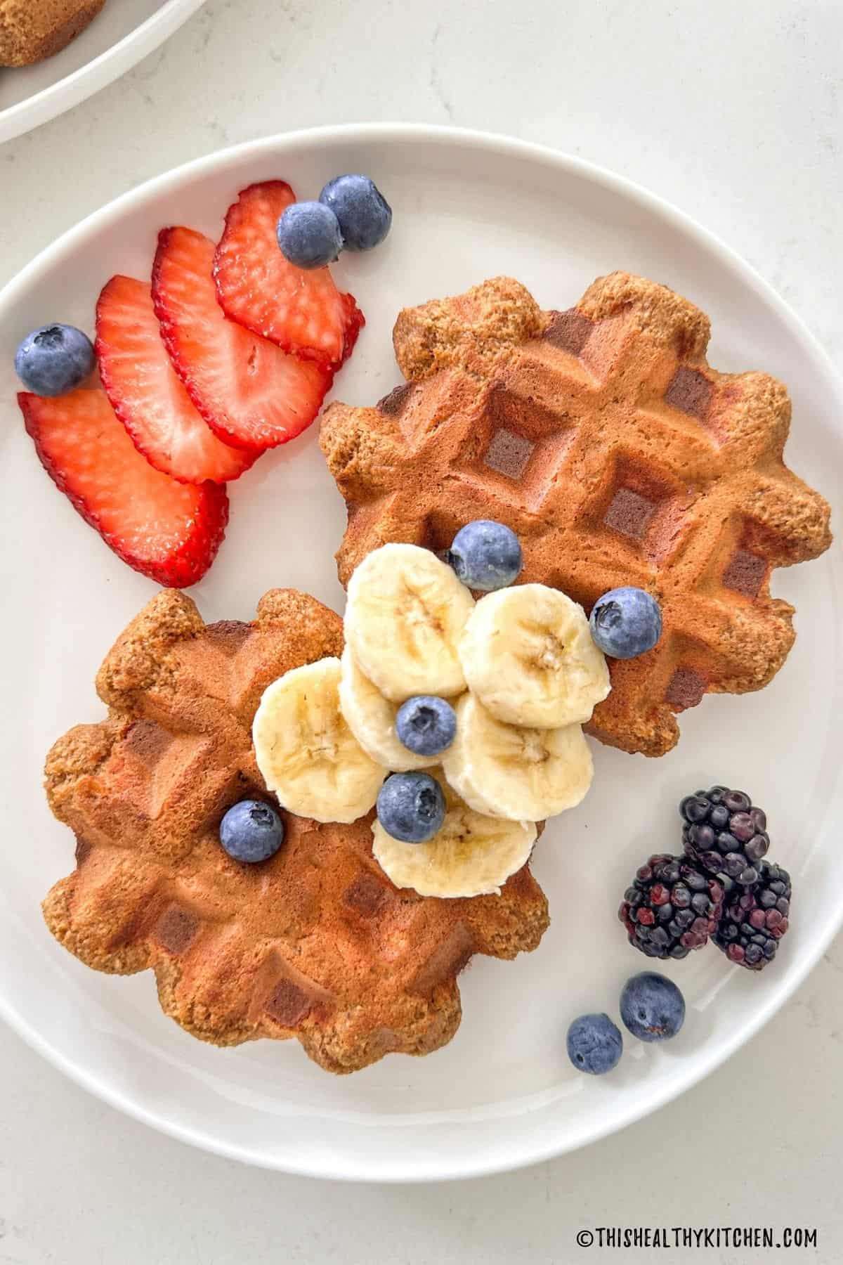 White plate with two banana protein waffles on it, with sliced bananas, strawberries and blueberries on top.